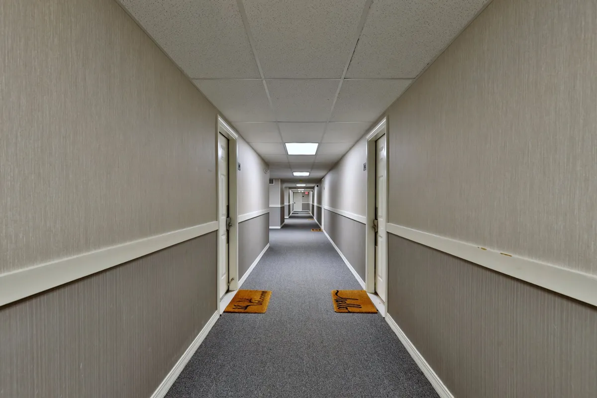 This image features a corridor with doors, carpet, and doormats, evoking the shared lobby or hallway entry. It captures the anticipation and transition as guests enter or move through the building’s common spaces.