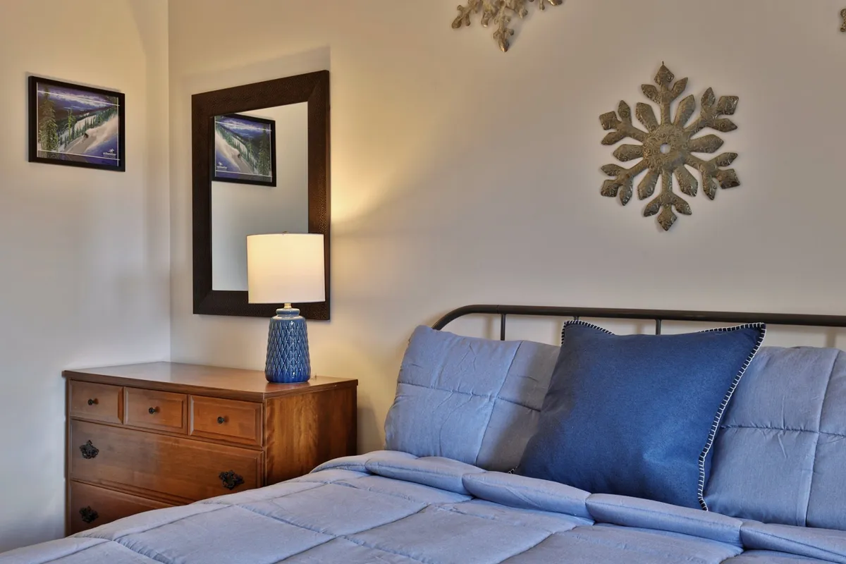 A serene bedroom featuring a queen bed with blue bedding, a wooden dresser, a decorative mirror, a lamp with a blue base, and snowflake wall decor. The overall mood is tranquil, with soft lighting and inviting textures creating a peaceful environment.