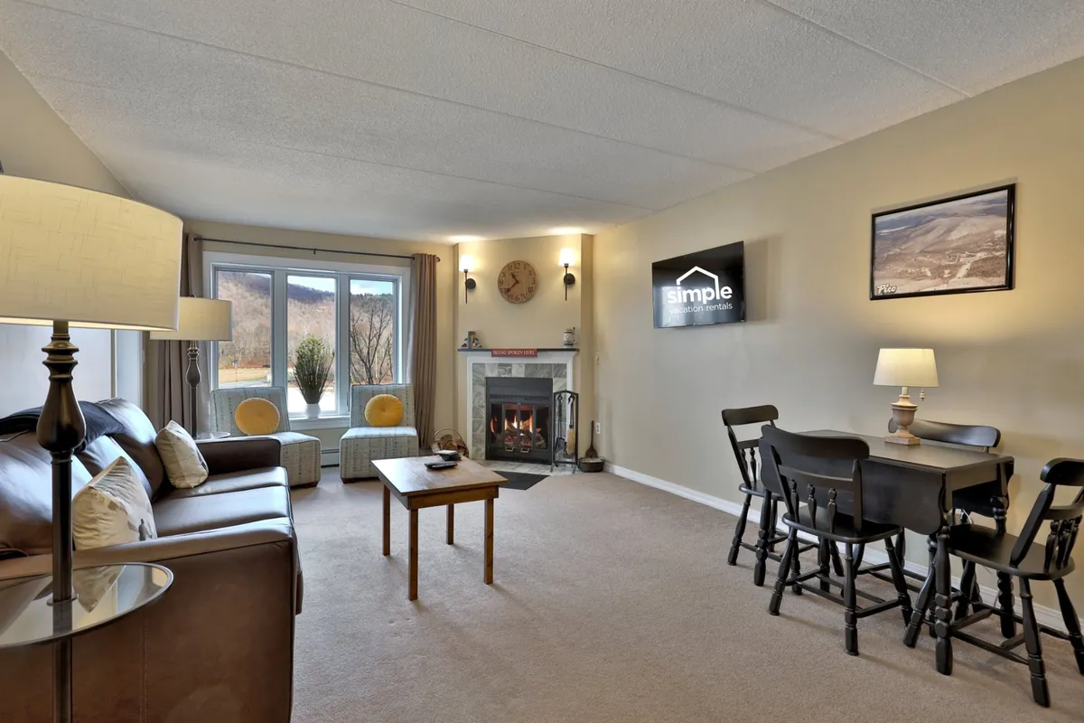 A welcoming living and dining area showcases a comfortable sofa positioned near a glowing fireplace, a dining table set for meals, and expansive windows that bring in natural light and hint at scenic views. The layout feels open and inviting, perfect for relaxing or gathering after a day outside.