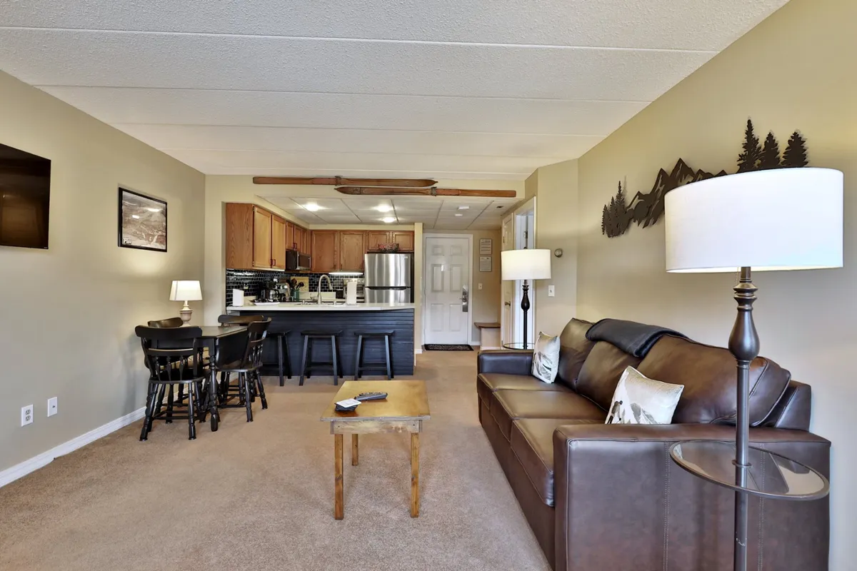 This image highlights a leather sofa and a wooden table adjacent to a bar seating area and kitchen. The cozy arrangement signals a warm, sociable environment, inviting for both relaxation and entertaining. The kitchen's visibility reinforces the connection between cooking, dining, and socializing.