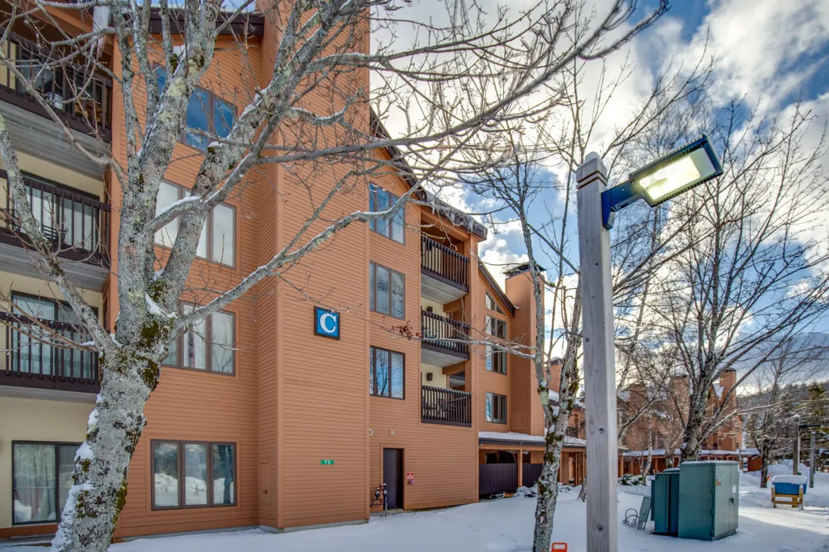 An exterior winter scene with the building's facade, snow-covered ground, bare trees, and a glowing streetlight, creating a cozy, inviting winter atmosphere. The image invites the viewer to imagine winter activities and the anticipation of returning to a warm, welcoming base after a day on the slopes.