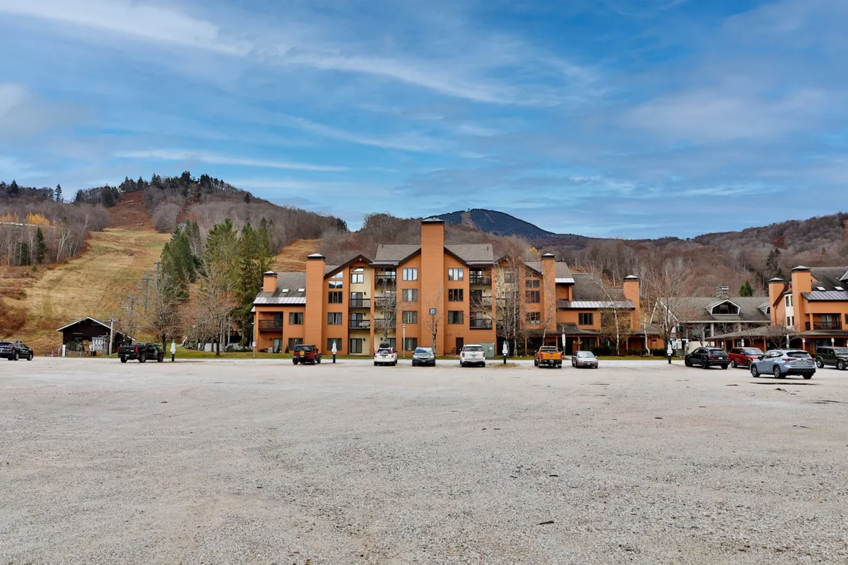 A wide exterior view of the main building set against the surrounding mountains and parking lot. The shot conveys a sense of arrival and orientation, highlighting the property's place in the Vermont mountain landscape, ideal for outdoor activities like skiing and hiking.
