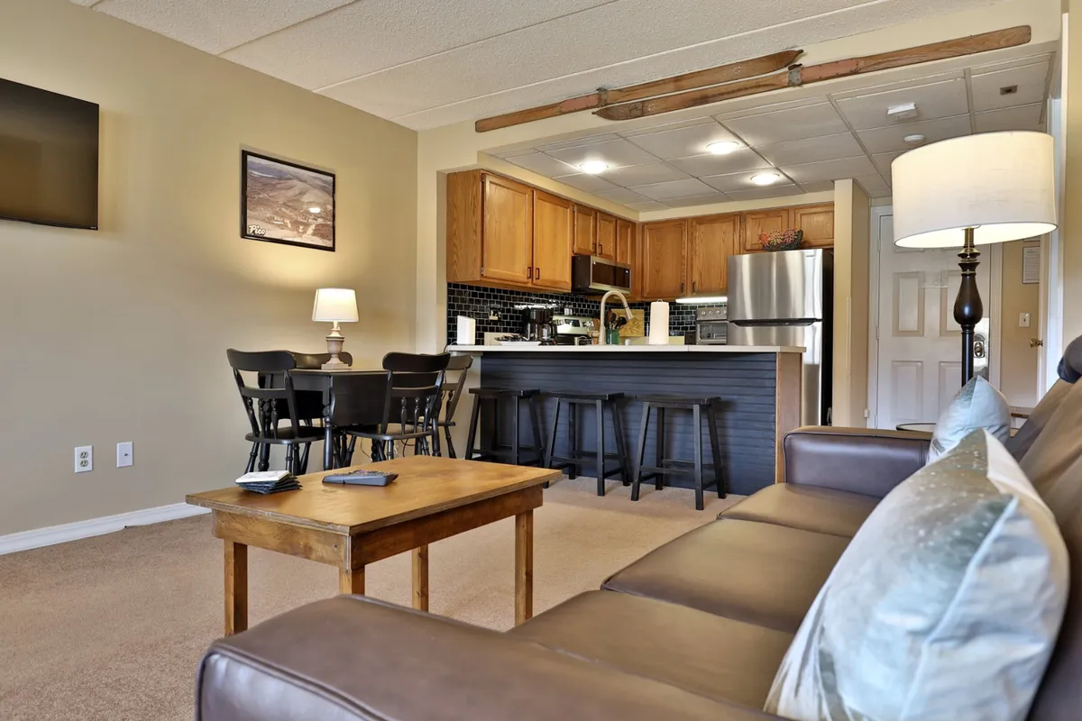 This image features a welcoming open-concept living and dining space, with a cozy sofa, a dining table set for group meals, a kitchen counter, and a softly lit lamp. The layout indicates a communal atmosphere, perfect for family gatherings, shared meals, and relaxed evenings at home.