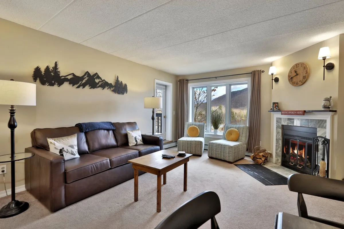 The image showcases a cozy living area featuring a plush sofa, a glowing fireplace, tasteful mountain-inspired wall art, and a large window revealing a scenic view—likely of the surrounding slopes or forest. The space invites guests to relax, socialize, or unwind after a day on the mountain.