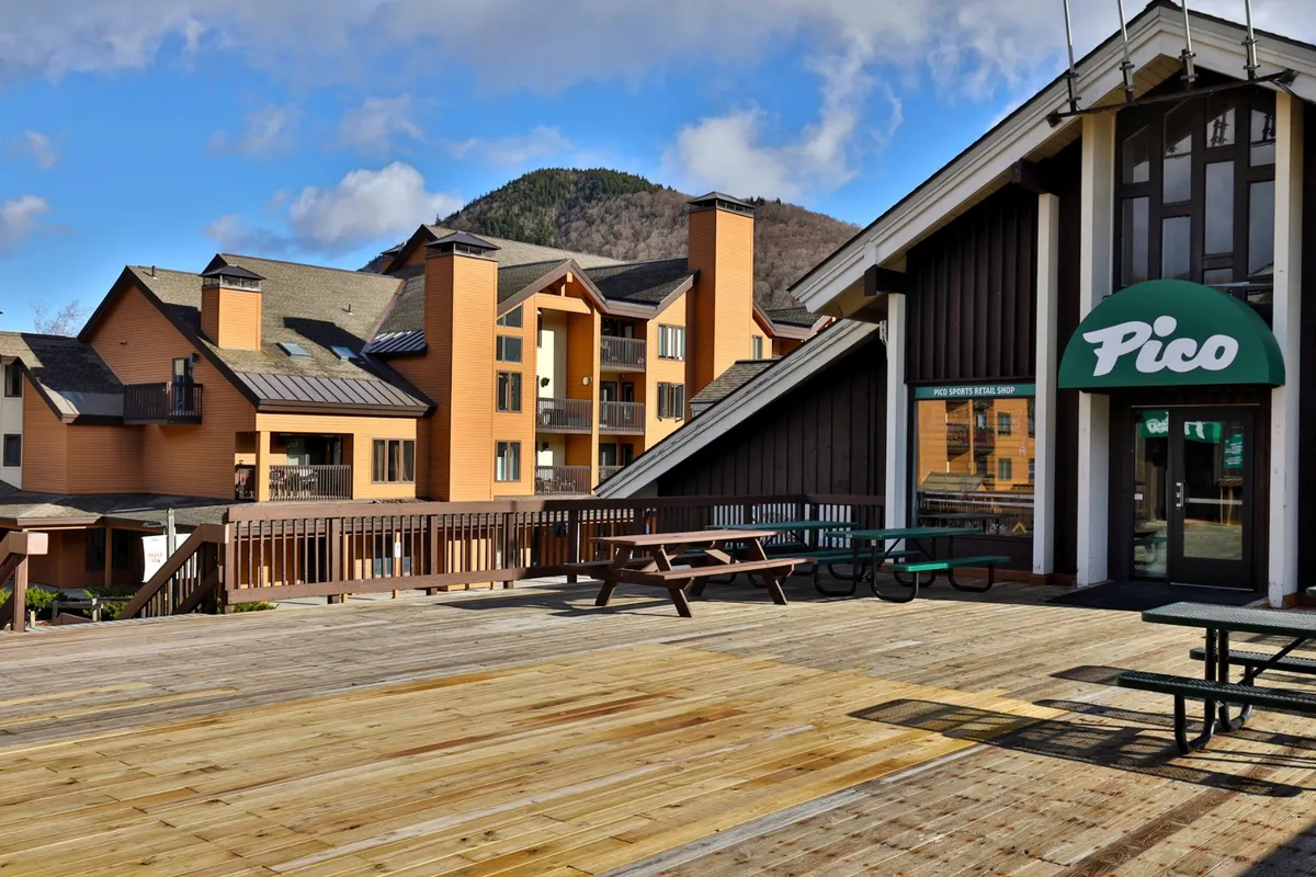 A welcoming exterior shot featuring the Pico sign, a wooden deck, and the mountain range as a backdrop. The image captures the inviting approach to the property and provides a clear connection to the mountain experience, with subtle hints of relaxation and social activity.