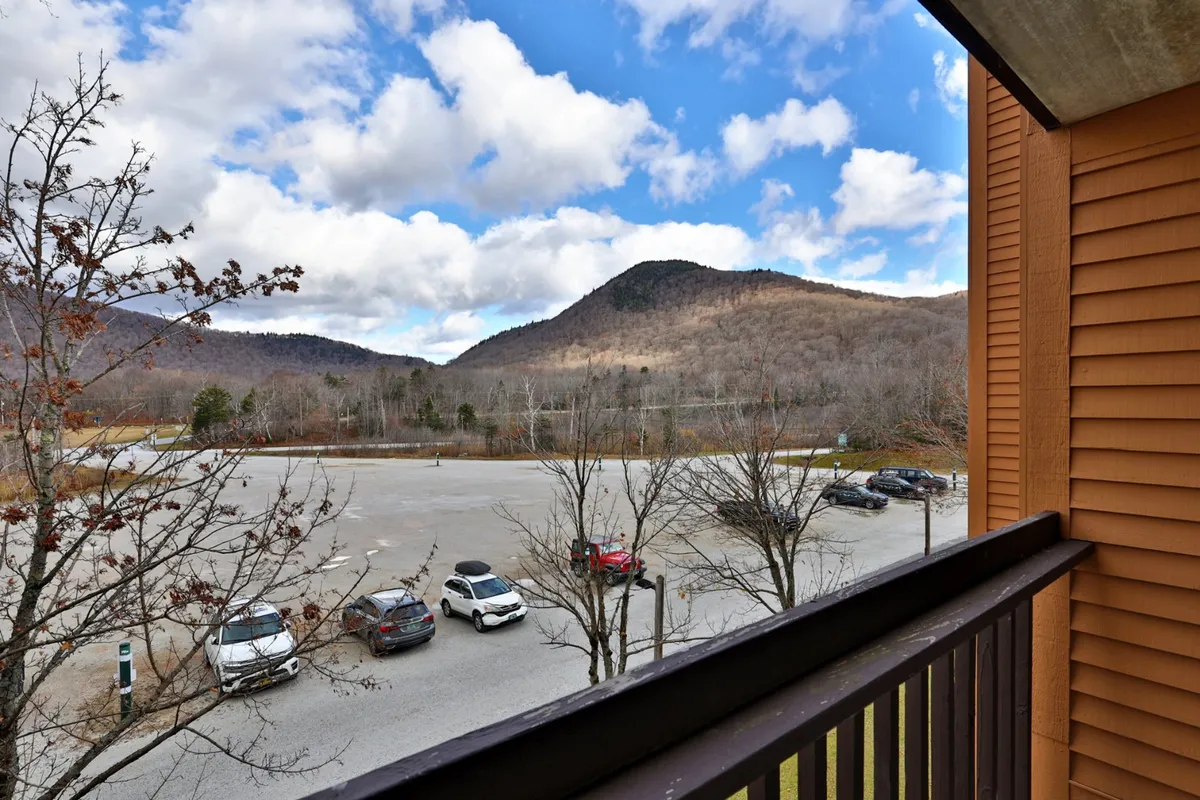 A sweeping view of majestic mountains under a dramatic sky, with trees framing the scene and a parking lot visible in the foreground. The natural landscape dominates the image, capturing the beauty and tranquility of the mountain environment.