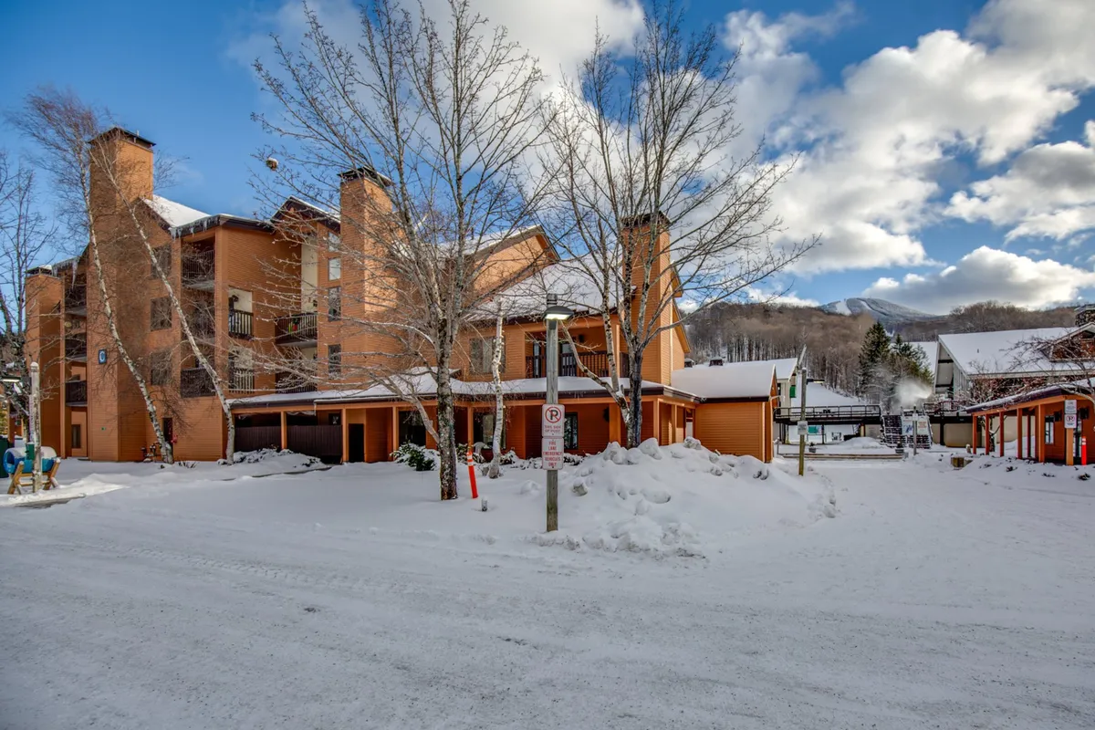 The exterior showcases the orange building façade against a snow-covered ground, with bare trees and mountains in the backdrop. The scene is lively and active, with visible ski and snowboard activities.