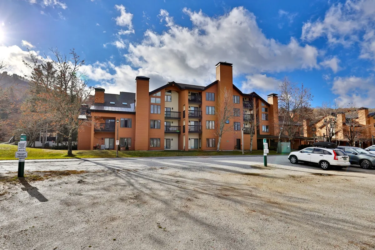 A multi-story orange building surrounded by mature trees and an open parking area. The setting radiates a welcoming, resort-like atmosphere, with expansive outdoor space hinting at opportunities for outdoor activities and gatherings. The building’s distinctive color and proximity to nature position it as a hub for both relaxation and adventure.